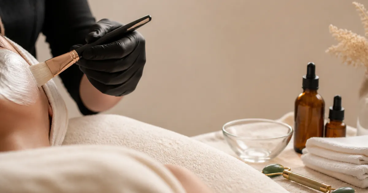 An esthetician's gloved hands brushing a cream mask onto a client during a facial, beside amber skincare bottles, folded towels and a jade roller